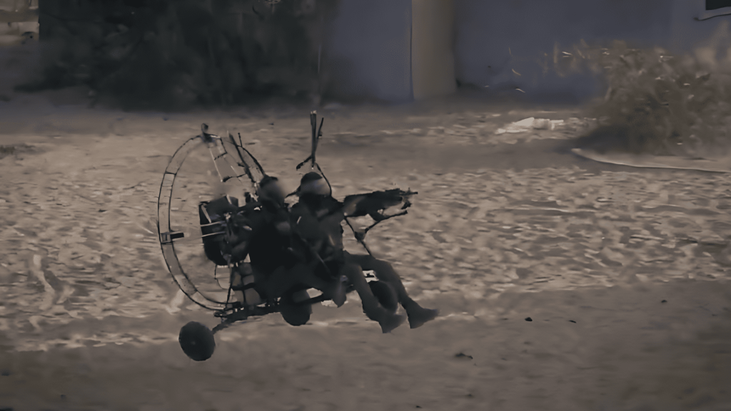 Paramotor armado con dos figuras volando bajo sobre terreno arenoso.