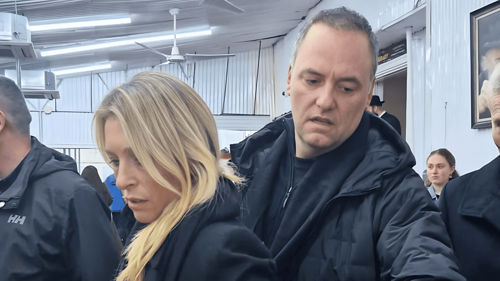 Mujer rubia y hombre con expresión seria en interior con gente.