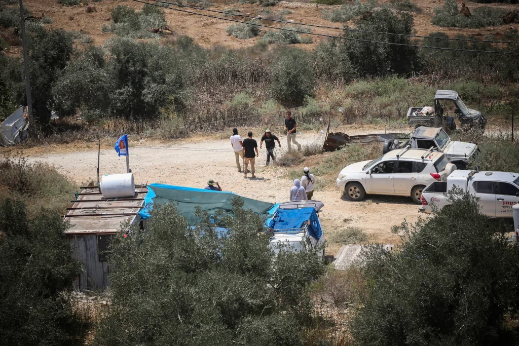 Hombres, vehículos, asentamiento informal en paisaje árido con bandera israelí.