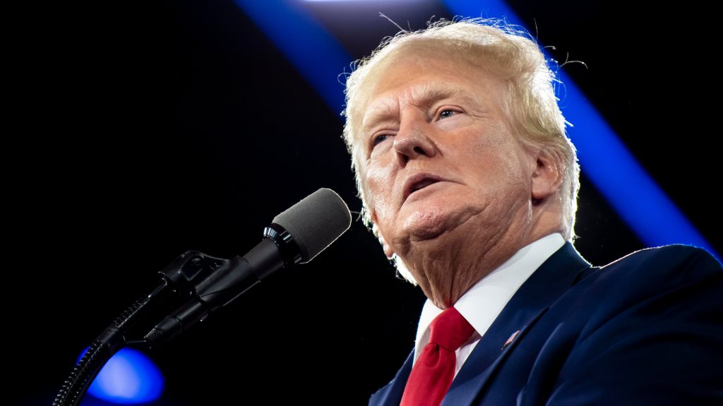 Donald Trump hablando en un escenario iluminado por luz azul.