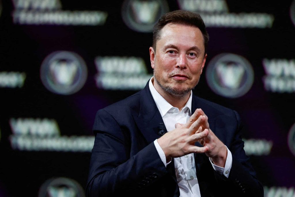 Elon Musk en evento, manos entrelazadas, mirando a cámara.