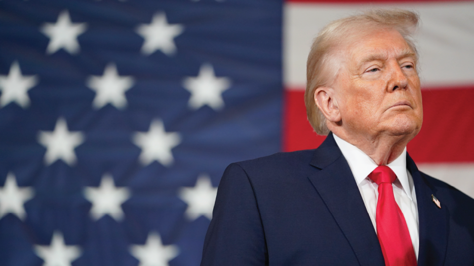 Donald Trump con traje azul y corbata roja frente a bandera de EE. UU.