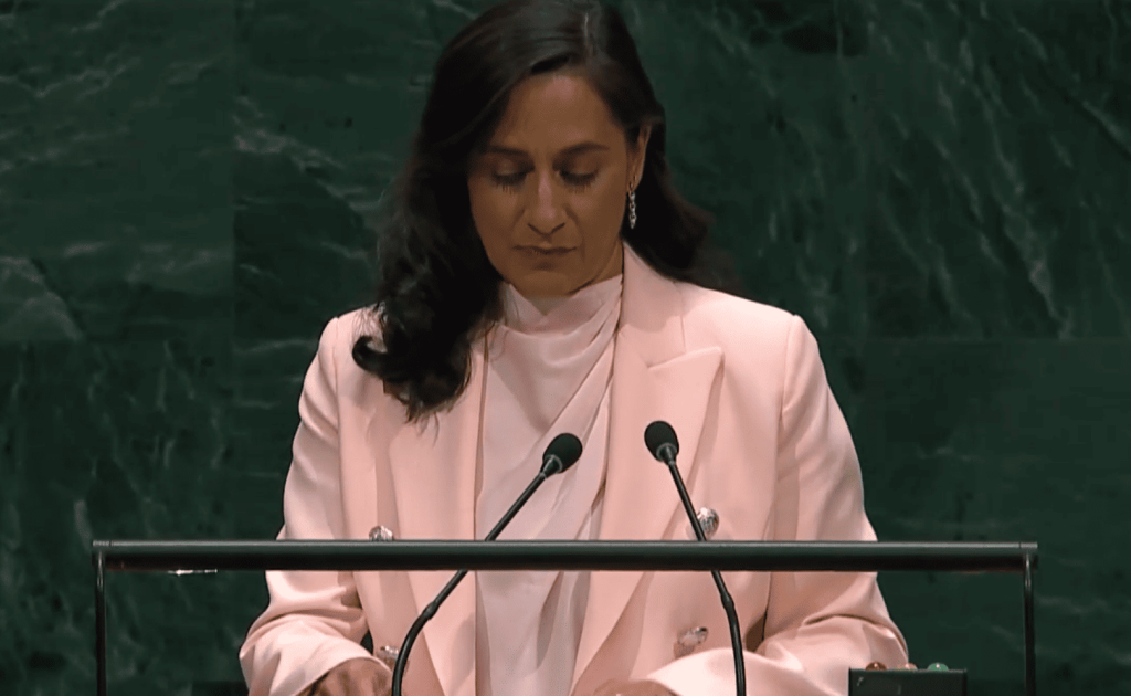 Mujer en podio, blazer rosa, mirando abajo, conferencia oficial.