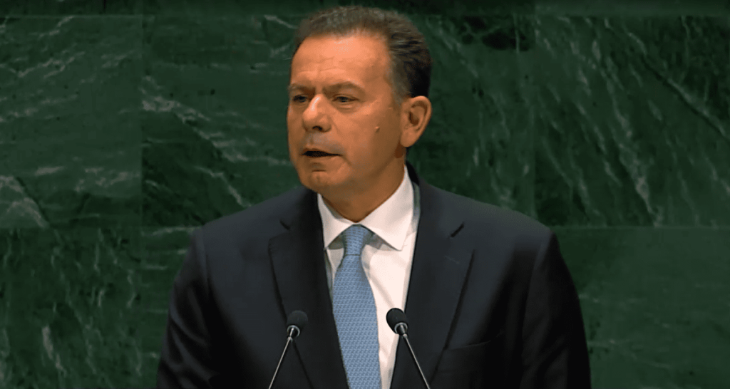 Hombre hablando en podio, traje, corbata, fondo verde mármol.