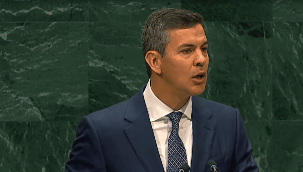 Hombre en traje hablando en podio, fondo verde marmolado.