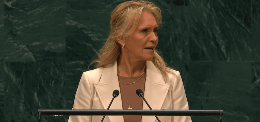 Mujer rubia en blazer blanco habla en podio con micrófonos.