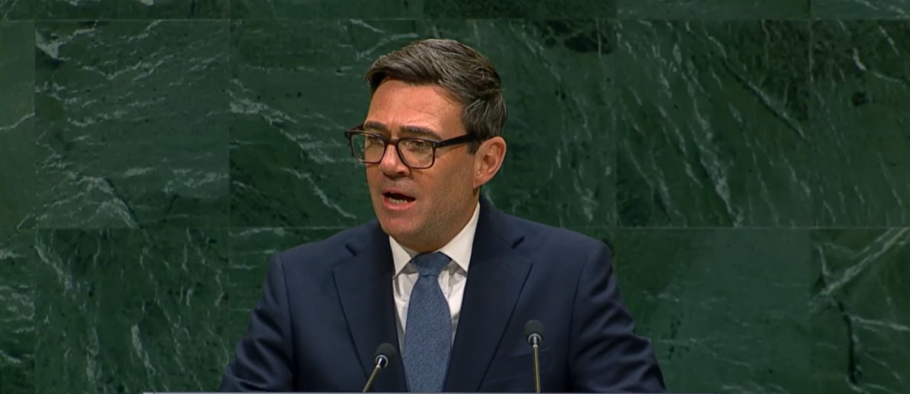 Hombre con gafas hablando en podio contra fondo verde mármol.