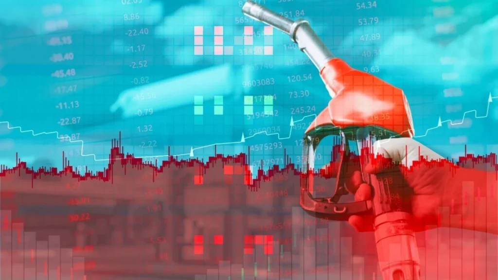 Mano sujetando manguera de gasolina sobre gráficos financieros rojos.