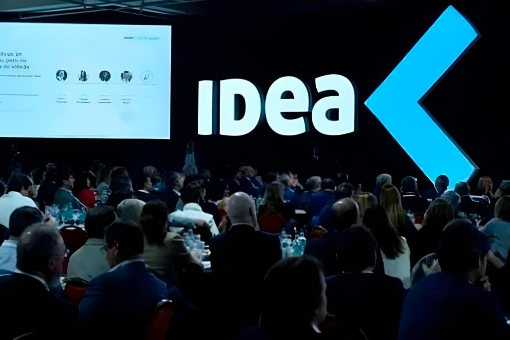 Público en un evento con logo 'IDea' y pantalla de presentación.