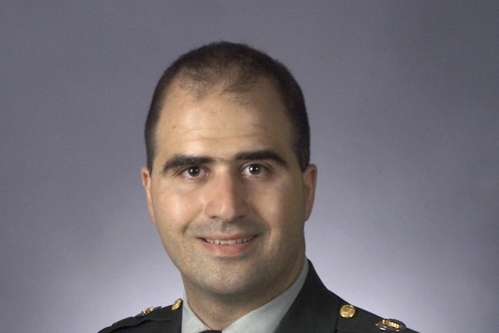 Hombre en uniforme militar sonriendo con fondo gris.