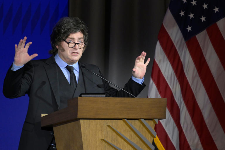 Javier Milei discursando enérgicamente en un atril frente a la bandera de EE. UU.