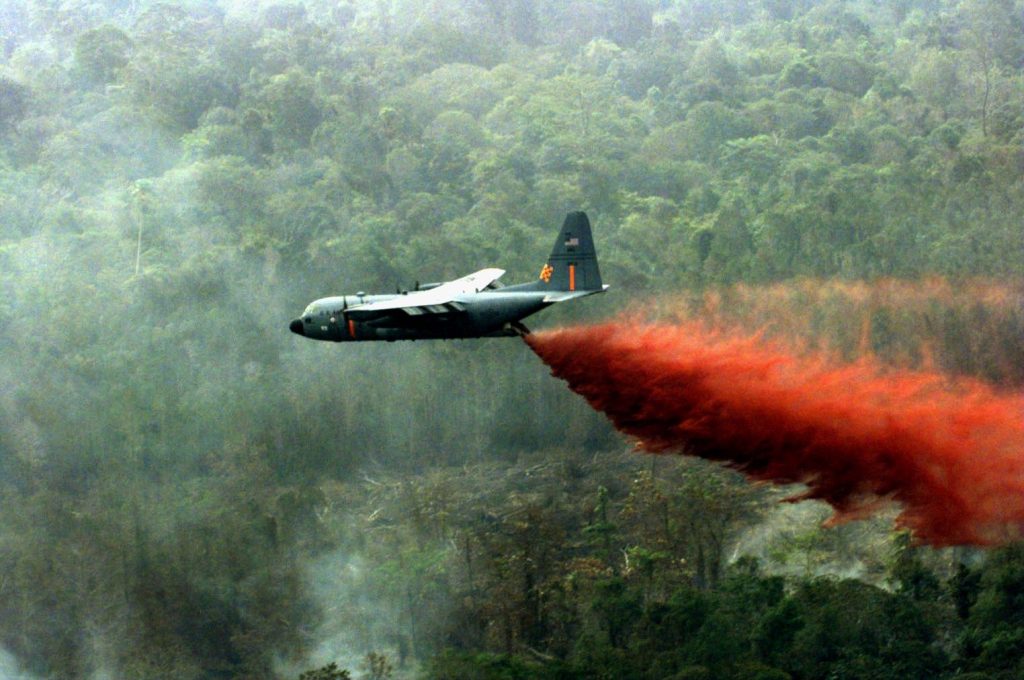 Avión militar dispersando sustancia roja sobre densa jungla brumosa.